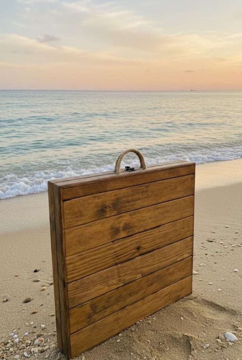 May include: A wooden folding table with a rope handle stands on a sandy beach. The table is constructed of horizontal wooden planks, stained a warm brown. The background features the ocean and a sunset.