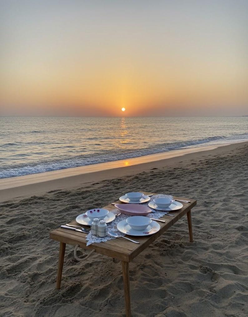 May include: A wooden picnic table set on a sandy beach, with a sunset over the ocean in the background. The table is set with white plates, bowls, silverware, and salt and pepper shakers. A pink serving dish is in the center.