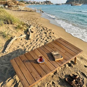 May include: A wooden table with a cup of tea, a book, and sandals on a sandy beach. The table is made of wooden planks and has four legs. The tea is in a small glass cup on a saucer. The beach has a view of the ocean and boats.
