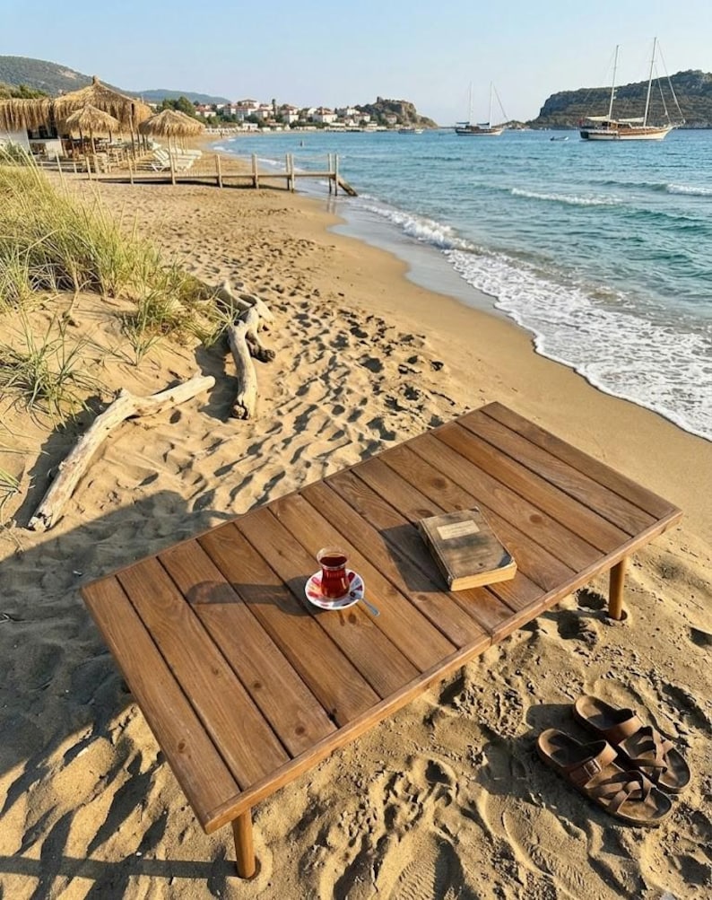 May include: A wooden table with a cup of tea, a book, and sandals on a sandy beach. The table is made of wooden planks and has four legs. The tea is in a small glass cup on a saucer. The beach has a view of the ocean and boats.