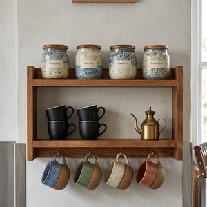 Könnte beinhalten: Ein zweistöckiges Wandregal aus Holz mit vier gesprenkelten Keramikdosen mit Holzdeckeln, beschriftet mit Darjeeling, Earl Grey, Green Tea und Herbal Blend. Darunter stehen vier schwarze Tassen, eine Messing-Teekanne und vier hängende Tassen in Blau, Grün, Creme und Terrakotta.