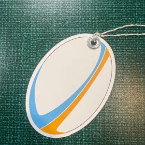May include: White oval tag with a blue and orange curved line design. The tag has a hole punched in the top with a silver metal grommet.