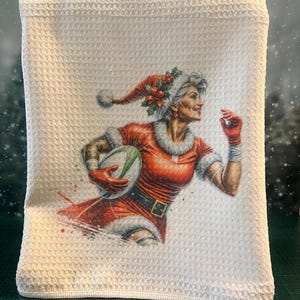 May include: White waffle weave kitchen towel featuring a printed image of a woman dressed as Santa Claus holding a rugby ball. She is wearing a red and white Santa suit with a green holly wreath on her hat. The background is a snowy scene.