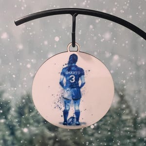 May include: A white round wooden ornament with a watercolor image of a person wearing a blue and white jersey with the number 3 on the back. The person is holding a rugby ball.