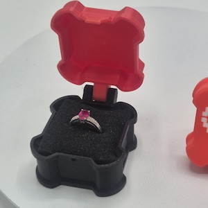Op de afbeelding: Een rode en zwarte ringdoos met een ring erin. De ring heeft een roze edelsteen. Een tweede rode doos met een gepixeld hartontwerp is ook aanwezig. De ringdoos is open en toont de ring.