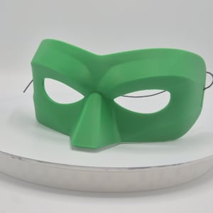 Op de afbeelding: Een effen groene masker met oogopeningen en een opvallende neus. Het masker is gemaakt van een glad materiaal en heeft een gebogen vorm. Zwarte touwtjes zijn bevestigd om te dragen. Geschikt voor kostuumfeesten of theatervoorstellingen.