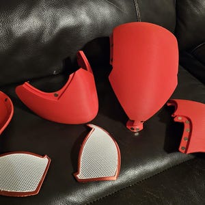 May include: Red 3D-printed mask components, including a face shield, cheek pieces, and eye covers. The mask parts are arranged on a dark leather surface. The eye covers have a white mesh design.