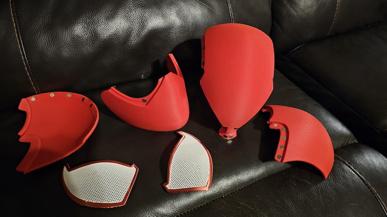 May include: Red 3D-printed mask components, including a face shield, cheek pieces, and eye covers. The mask parts are arranged on a dark leather surface. The eye covers have a white mesh design.