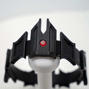 May include: A black crown featuring a series of angular, geometric panels and a single, small, hexagonal red gem centered on the front-most panel. The crown has a rigid, structured design with pointed elements extending upward along the top edge.