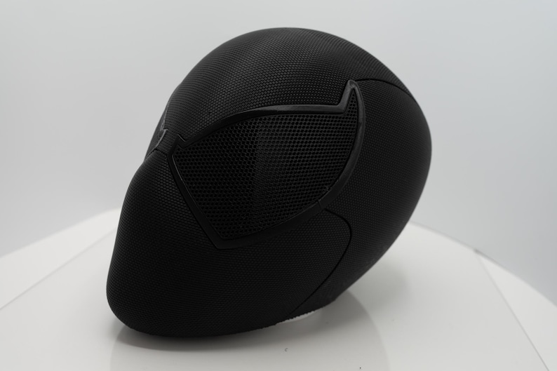 May include: A black mask with a textured surface and mesh eye covering. The mask has a streamlined design, with a smooth, curved shape. The eye covering is a darker black, creating contrast. Likely for cosplay or costume purposes.