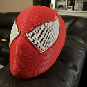 May include: Red and white Spider-Man mask with a textured surface. The mask has white mesh eye coverings with a copper-coloured trim. The mask is resting on a black leather sofa.