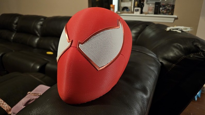 May include: Red and white Spider-Man mask with a textured surface. The mask has white mesh eye coverings with a copper-coloured trim. The mask is resting on a black leather sofa.