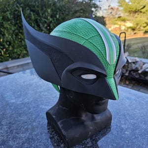 May include: A black and green superhero mask with pointed ears, displayed on a black mannequin head. The mask has a green textured surface with a white stripe and eye mesh. Likely for cosplay or costume use.