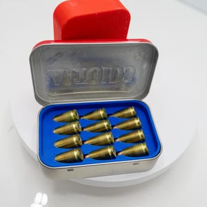 Pode incluir: Uma lata de metal aberta com tampa vermelha, revelando um interior azul contendo doze objetos de cor latão. A tampa da lata tem a palavra "VINTAGE" em relevo. Os objetos estão cuidadosamente dispostos dentro.