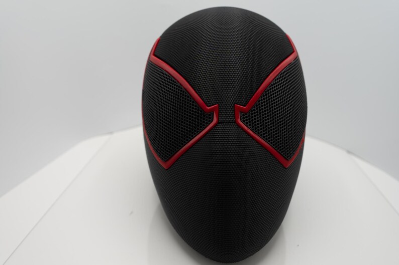 May include: A black mask with red detailing, with a mesh pattern over the eye areas. The mask has a smooth, curved form and is likely a costume or cosplay item.