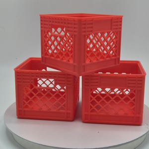 May include: Three small, red plastic crates stacked in a pyramid formation. Each crate has a square shape with a lattice design on the sides. The crates are sitting on a white surface.