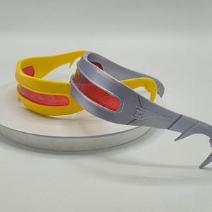 Może przedstawiać: Dwie pary okularów w stylu superbohatera. Jedna para jest żółta z czerwoną soczewką, a druga szara z czerwoną soczewką. Okulary mają zakrzywioną konstrukcję i prawdopodobnie są przeznaczone do cosplayu lub kostiumów.