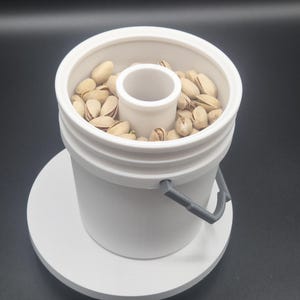 Op de afbeelding: Een witte plastic pistachekom met een centrale cilinder en een grijze handgreep. De kom is gevuld met pistachenoten. De kom staat op een witte cirkelvormige basis.