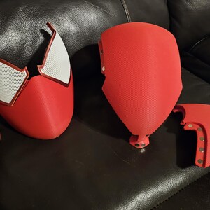May include: A red and white Spider-Man mask and costume components. The mask features white mesh eye sections and a red outline. Other red pieces include a chest plate and arm sections. The items are arranged on a dark surface.