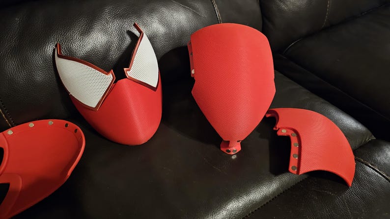 May include: A red and white Spider-Man mask and costume components. The mask features white mesh eye sections and a red outline. Other red pieces include a chest plate and arm sections. The items are arranged on a dark surface.