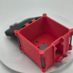 May include: A red, square-shaped container with a hinged lid. The container has a rectangular opening and is made of a hard plastic material. The container is sitting on a white surface.