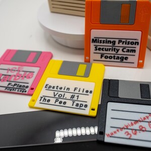 Könnte beinhalten: Eine Sammlung farbenfroher Disketten, jede mit einzigartigen Etiketten. Die orangefarbene Diskette trägt die Aufschrift "Missing Prison Security Cam Footage". Die rosa Diskette trägt die Aufschrift "ICE Barbie F-Plane Flight Logs". Die gelbe Diskette trägt die Aufschrift "Epstein Files Vol. #1 The Pee Tape."