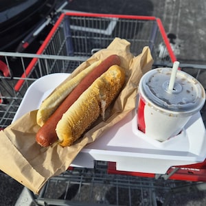 Może przedstawiać: Hot dog w bułce sezamowej i druga bułka, z napojem gazowanym w kubku ze słomką, na białej tacy. Taca znajduje się w wózku sklepowym.