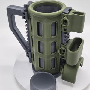 May include: A green and gray mug with a tactical design, featuring a cylindrical body with rectangular cutouts and a handle. The mug includes a lid and additional attachments, resembling military equipment.