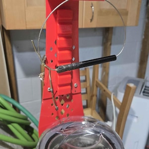 May include: A red metal stand with a wire loop and a black cylindrical component. Below, a gray and clear device with the word "SILCA" is visible. The stand has a ladder-like design and is set against a wooden cabinet.