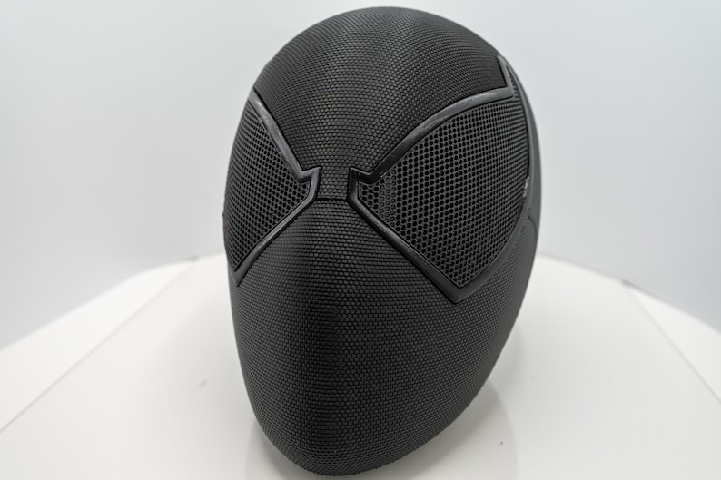 May include: A black mask with a textured surface and mesh eye coverings. The mask has a distinct design with angular shapes around the eyes, and is likely a costume or cosplay accessory.