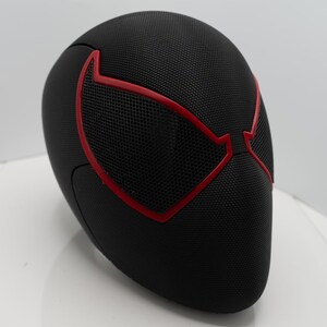 May include: Black mask with red detailing, featuring a mesh eye design. The mask has a textured surface, possibly for a costume or collectible. The red outlines the eye shapes, creating a striking contrast against the black.