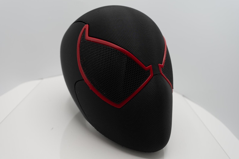 May include: Black mask with red detailing, featuring a mesh eye design. The mask has a textured surface, possibly for a costume or collectible. The red outlines the eye shapes, creating a striking contrast against the black.
