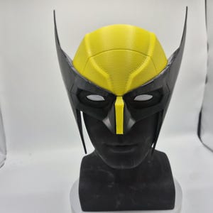 Puede incluir: Una máscara de Wolverine negra y amarilla, con una pieza para la cabeza amarilla y detalles negros para los ojos y la cara. La máscara tiene extensiones puntiagudas en forma de orejas y se muestra en un busto negro.
