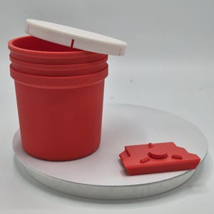 Op de afbeelding: Een rode container met een witte deksel en een apart rood stuk. De container is cilindrisch met een geribbelde buitenkant. De deksel is rechthoekig. Het aparte stuk is een kleine, rode, geometrische vorm.
