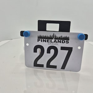 May include: A white race bib displaying the number 227 in black. The bib features the text "PINELANDS GRAVEL" and a tree graphic. It is held by a black bracket with blue knobs.