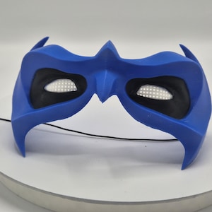 Può includere: Una maschera da supereroe blu con copriocchi neri e inserti per occhi in rete bianca. La maschera ha una parte superiore a punta ed estensioni laterali. Un sottile filo nero si estende dalla maschera.