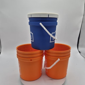 以下が含まれることがあります： 3つのプラスチックバケツ。白いハンドルと蓋が付いた青いバケツが、2つのオレンジ色のバケツの上に積み重ねられています。青いバケツには「Lowe's」と書かれた白い文字があります。オレンジ色のバケツにも白い文字があります。