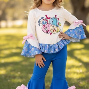 May include: A cream-colored long-sleeved top with a heart-shaped graphic and the text "FLORES DAY". The top has blue floral print bell sleeves with pink bows. The outfit includes blue bell-bottom pants with pink bows.