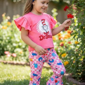 May include: Pink Dr. Seuss themed outfit for girls. The set includes a pink t-shirt with ruffle sleeves and a graphic of the Cat in the Hat, and matching pink checkered pants with a flared leg. A blue headband completes the look.