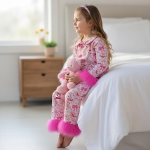 以下が含まれることがあります： ピンクのパジャマセット、お揃いのヘアバンドとぬいぐるみ付き。パジャマは、リボンやその他のデザインの繰り返し模様が特徴です。パジャマパンツは、足首に明るいピンクのフェイクファーのトリムが付いています。