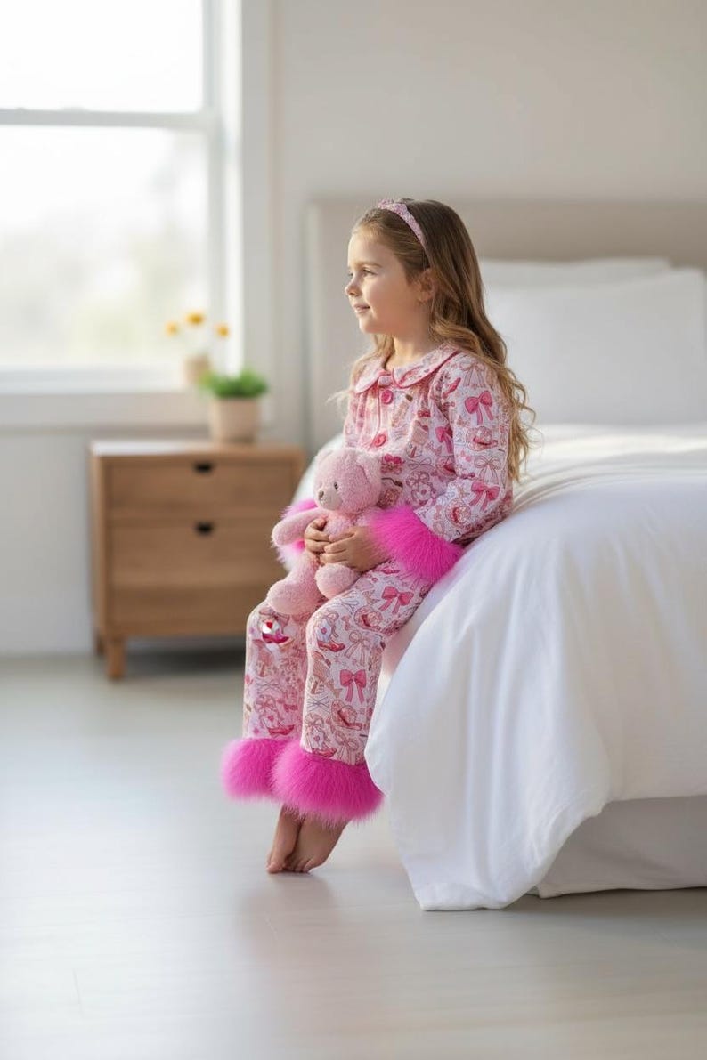 以下が含まれることがあります： ピンクのパジャマセット、お揃いのヘアバンドとぬいぐるみ付き。パジャマは、リボンやその他のデザインの繰り返し模様が特徴です。パジャマパンツは、足首に明るいピンクのフェイクファーのトリムが付いています。