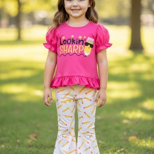 May include: A young girl wears a bright pink top with the words "Lookin' Sharp" and a pencil graphic, paired with white pants patterned with yellow pencils. The top has ruffled sleeves and a ruffled hem. The pants are flared at the bottom.