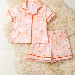 Peut inclure: Ensemble de pyjama rose avec un motif de lapin et de carotte. Le haut &agrave; manches courtes a une fermeture boutonn&eacute;e et un col, avec une bordure orange. Un short assorti compl&egrave;te l'ensemble. Le pyjama est en mati&egrave;re douce.