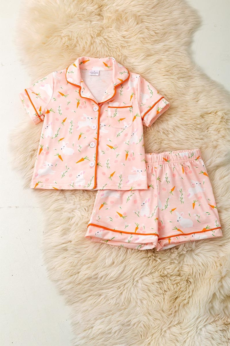 Peut inclure: Ensemble de pyjama rose avec un motif de lapin et de carotte. Le haut &agrave; manches courtes a une fermeture boutonn&eacute;e et un col, avec une bordure orange. Un short assorti compl&egrave;te l'ensemble. Le pyjama est en mati&egrave;re douce.
