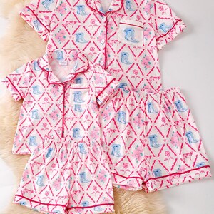 K&ouml;nnte beinhalten: Passende Pyjama-Sets in Wei&szlig; mit einem rosa-blauen Blumen- und Stiefelmuster. Jedes Set besteht aus einem kurz&auml;rmeligen Oberteil mit Kn&ouml;pfen und Shorts, die mit rosa Paspeln versehen sind. Die Sets werden auf einer flauschigen, cremefarbenen Oberfl&auml;che pr&auml;sentiert.