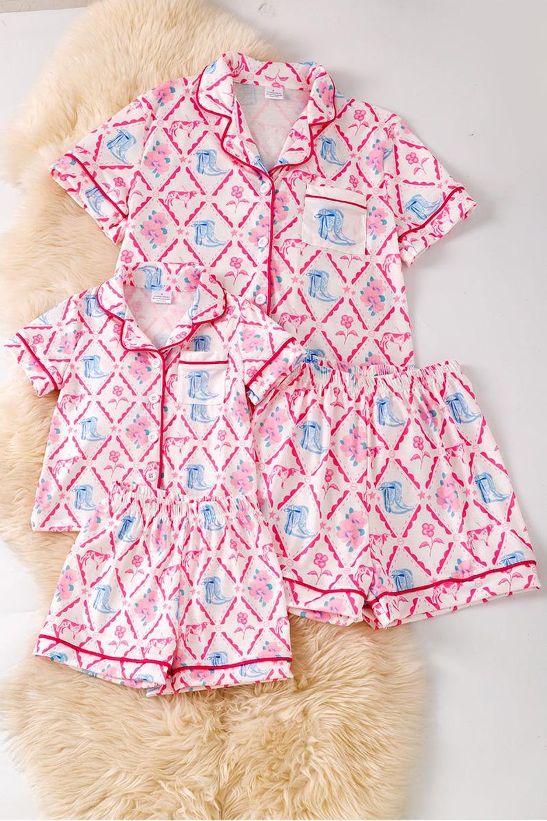 K&ouml;nnte beinhalten: Passende Pyjama-Sets in Wei&szlig; mit einem rosa-blauen Blumen- und Stiefelmuster. Jedes Set besteht aus einem kurz&auml;rmeligen Oberteil mit Kn&ouml;pfen und Shorts, die mit rosa Paspeln versehen sind. Die Sets werden auf einer flauschigen, cremefarbenen Oberfl&auml;che pr&auml;sentiert.