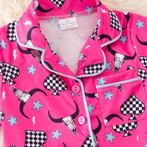 Pode incluir: Camisola de pijama rosa com um padr&atilde;o de espadas quadriculadas, caveira e estrelas. A parte superior tem um fecho de bot&atilde;o e um bolso do lado esquerdo.