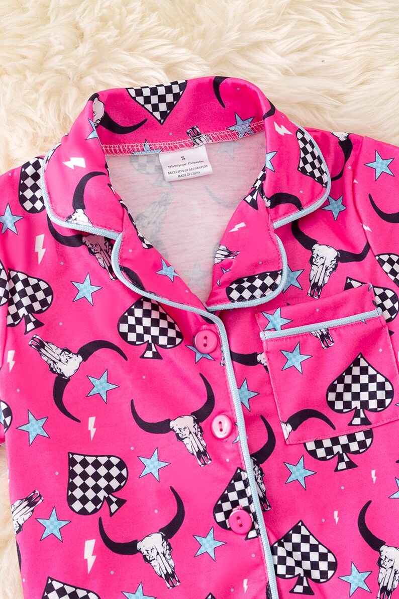 Pode incluir: Camisola de pijama rosa com um padr&atilde;o de espadas quadriculadas, caveira e estrelas. A parte superior tem um fecho de bot&atilde;o e um bolso do lado esquerdo.