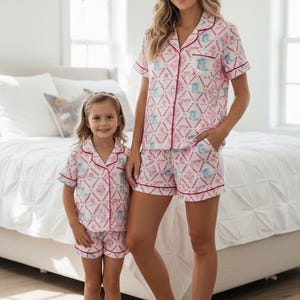 K&ouml;nnte beinhalten: Passende rosa-wei&szlig;e Pyjama-Sets mit einem sich wiederholenden Muster aus Stiefeln und floralen Designs. Die Sets umfassen kurz&auml;rmelige Hemden mit Kn&ouml;pfen und Shorts, die mit roten Paspeln versehen sind. Eine Mutter und ein Kind tragen die passenden Sets.
