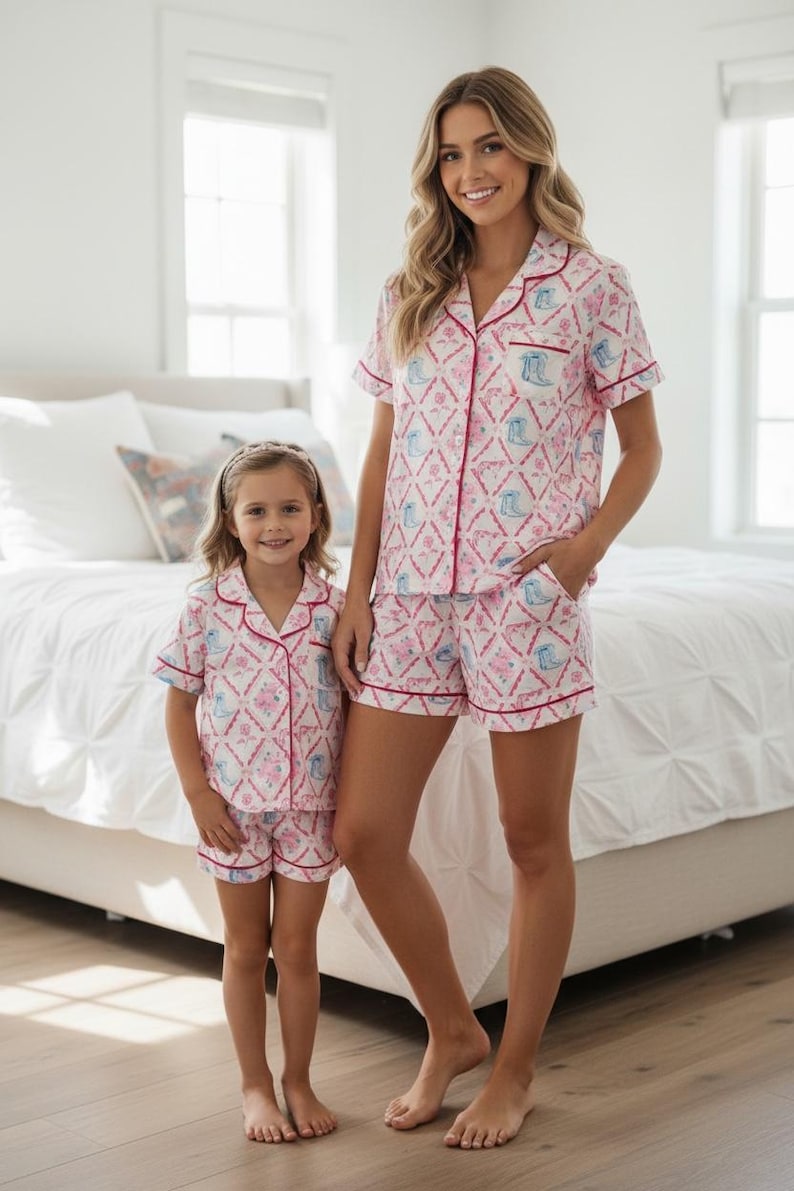 K&ouml;nnte beinhalten: Passende rosa-wei&szlig;e Pyjama-Sets mit einem sich wiederholenden Muster aus Stiefeln und floralen Designs. Die Sets umfassen kurz&auml;rmelige Hemden mit Kn&ouml;pfen und Shorts, die mit roten Paspeln versehen sind. Eine Mutter und ein Kind tragen die passenden Sets.